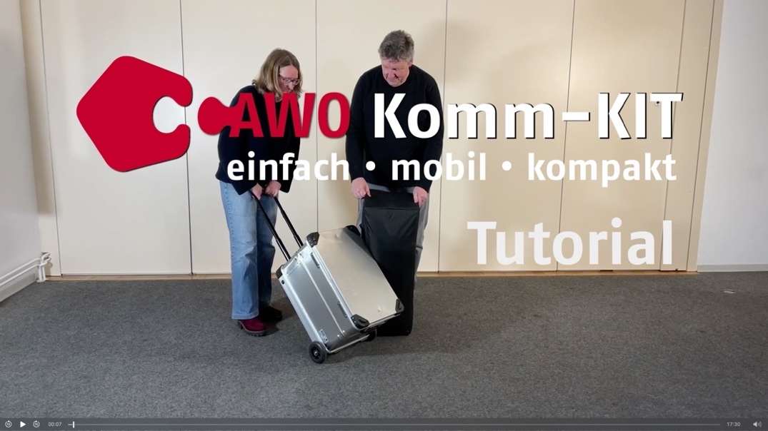 Screenshot eines Videos: Im Vordergrund AWO Komm-Kit - einfach, mobil, kompakt - Tutorial. Im Hintergrund eine Frau und ein Mann mittleren Alters in einem Raum. Die Frau hat einen Alurollkoffer in der Hand, der Mann hält eine schwarze, rechteckige Tasche, die auf dem Boden steht. Beide stehen nebeneinander und schauen auf den Koffer und die Tasche.